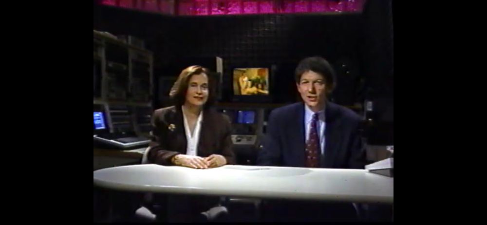a screenshot of two people in suits sit at a table and address the camera. behind them on a tv is a video of two people having sex