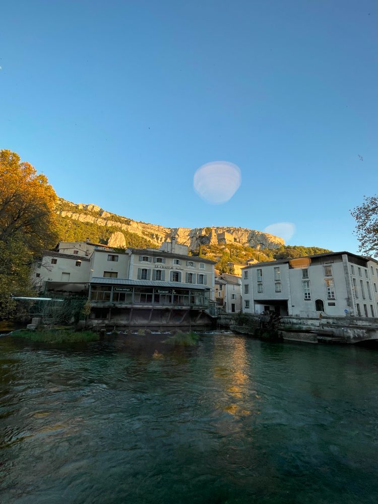Blick auf die hohe Felswand im Hintergrund des Ortes, im Vordergrund sieht man den Fluss, dahinter die Häuser des Ortes, die Felswand leuchtet in der Abendsonne und darüber blauer Himmel