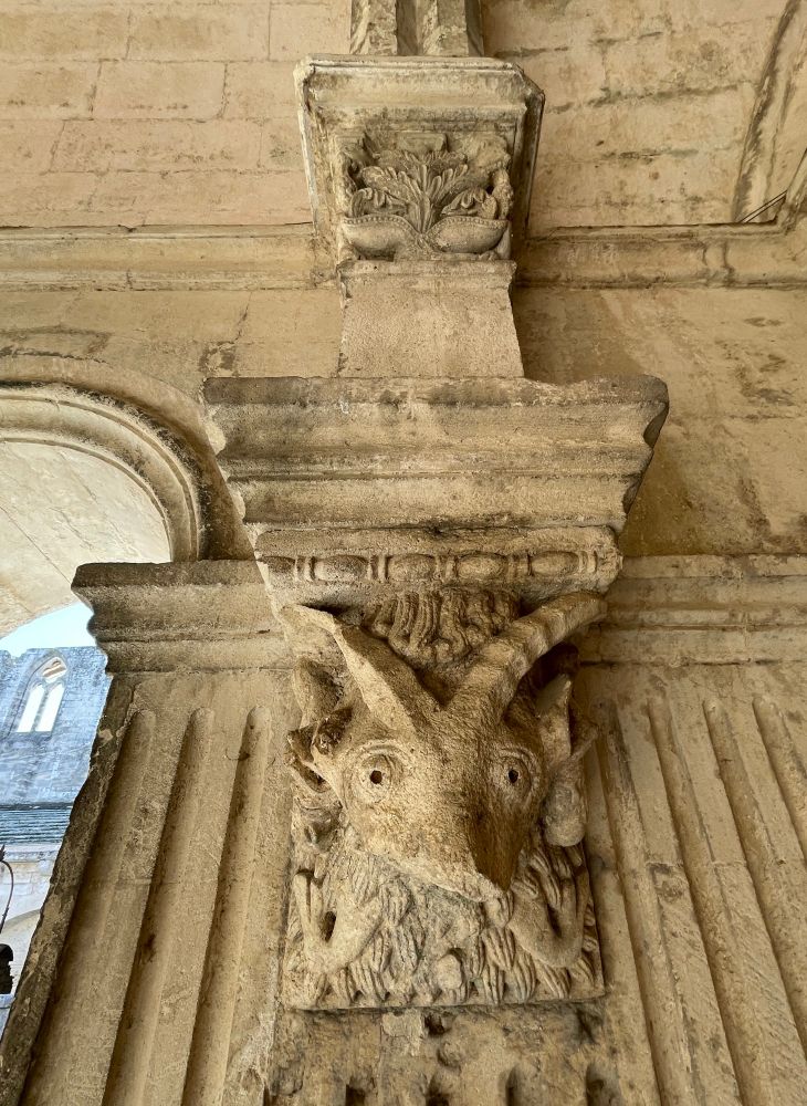 Ein Kapitell im Kreuzgang der Kirche St. Trophime in Arles zeigt eine ziemlich ulkige Gestalt, mir ist nicht ganz klar, ob es sich um einen Ziegenbock oder um einen menschlichen Kopf mit Ziegenhörnern handelt, vielleicht ein Teufel?! Das Gesicht ist nicht komplett erhalten oder irgendwie schief geraten, die Hörner sind leicht gewählt, riesige Augen schauen dich an, und unten scheint ein schuppiger Bart erkennbar zu sein. Zwischen den Hörnern sieht man wenige Formen, die vielleicht Haare darstellen, darüber ein waagerechter Schmuckstab.