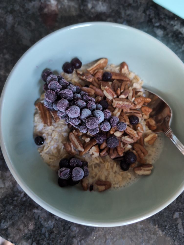 baby blue ceramic bowl of oatmeal with crushed pecans and frozen blueberries in. most of the blueberries are frozen together in a distinct corncob.