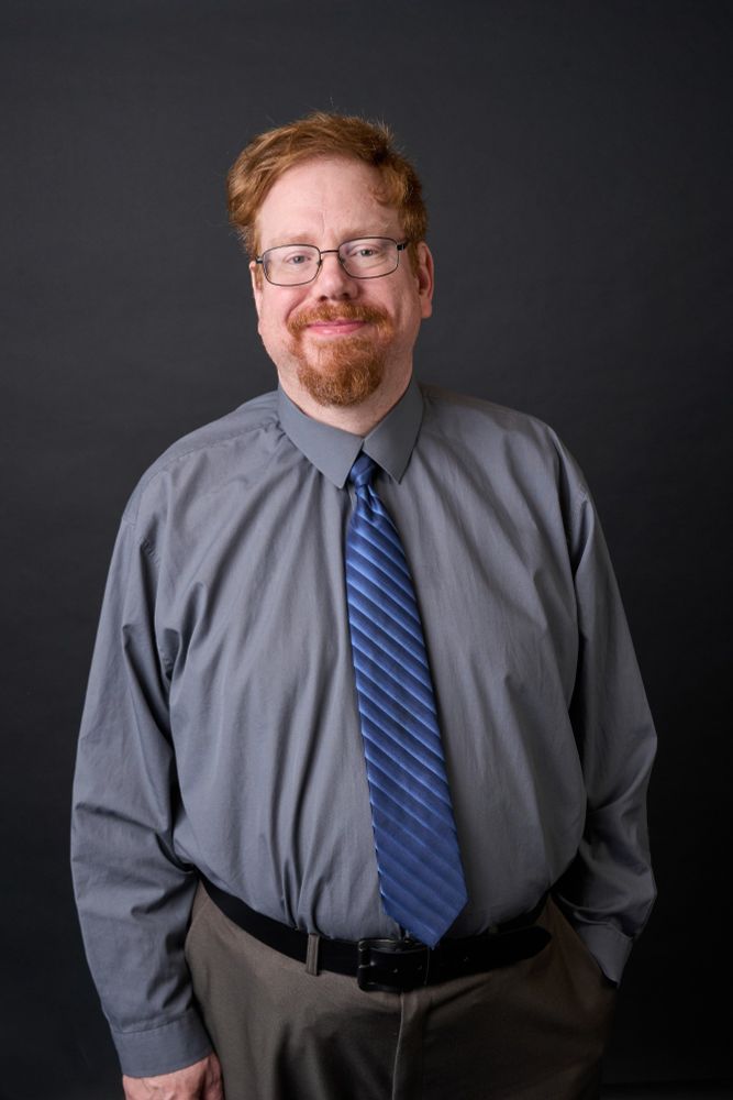 A photo of a red haired man is a grey shirt and blue necktie. 