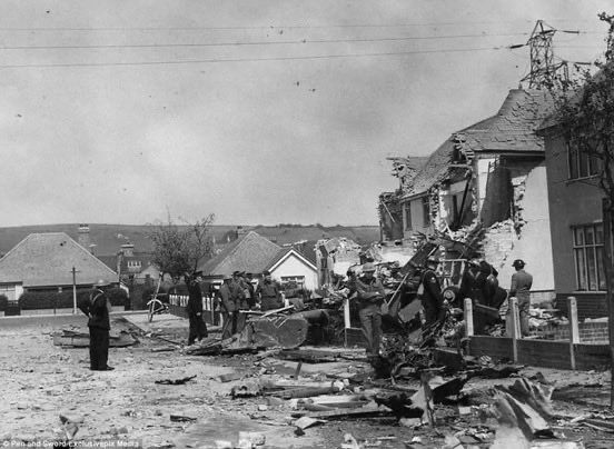 Suburban street littered with the wreckage of an aircraft which has crashed into two semi-detached houses. High-tension power lines are visible behind. Police, military and ARP personnel are in attendance.