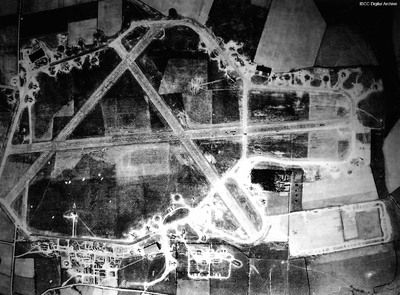Overhead photograph, taken from medium altitude, of a large airfield, with three long concrete runways arranged in an ‘A’ shape. Surrounding them is a long perimeter track with multiple circular dispersal bays for aircraft.