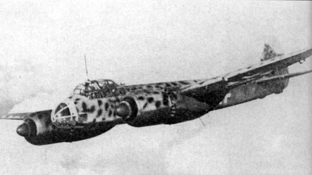 Junkers 88 with streamlined glazed nose and no ventral gondola flying above a broken cloud layer. It carries mottled camouflage above, with black undersides.