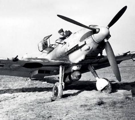 Messerschmitt 109 fighter on a French airfield, carrying a single 250kg bomb on an under fuselage rack. A Luftwaffe airman, or possibly the pilot, is checking something in the cockpit.