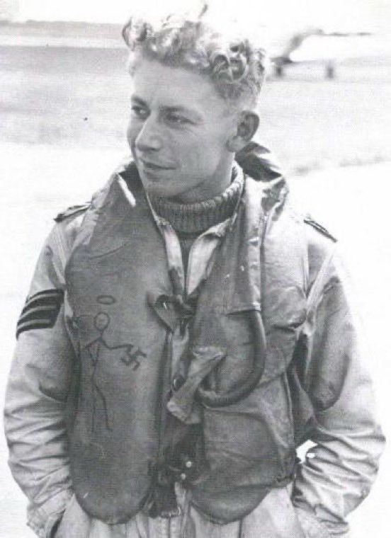 Curly haired sergeant pilot in light coloured flying overalls. He’s wearing a life jacket with a drawing of a stick figure with a halo, holding a swastika in one hand.