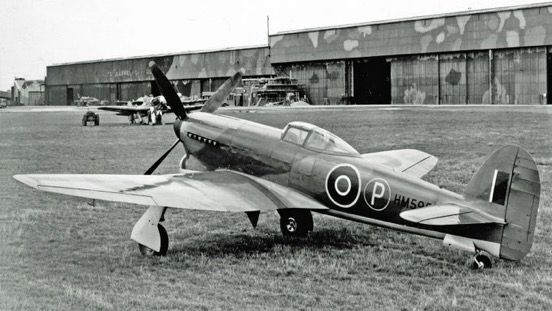 The first Tempest to fly was Mark V HM595, which shared the tailfin and original ‘car door’ cockpit of the Typhoon. These would change before the type entered service. 📷 baesystems.com (Single seater fighter with a four bladed propeller and highly polished, thin wing parked on grass in front of camouflaged hangars)