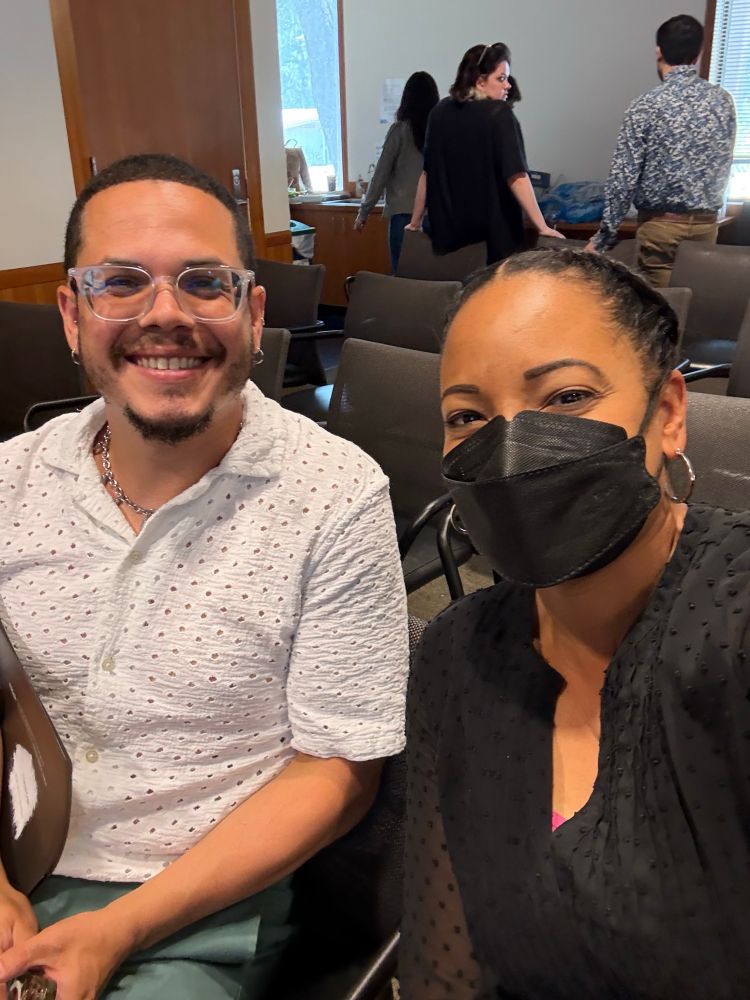 Two scientists sitting in a seminar room.
One is masked. They are both smiling. 