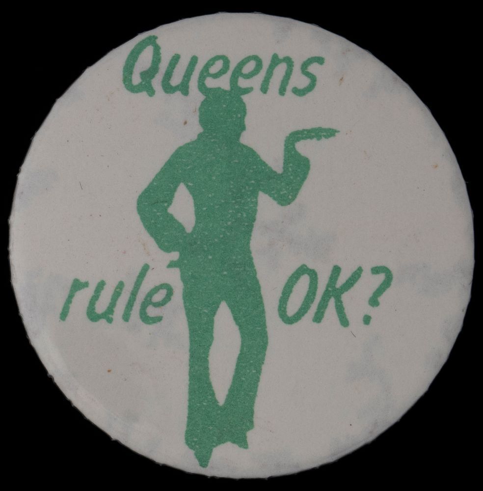 White badge pin with green silhouette of a limp-wristed foppish man with a hand on his hip reading “Queens rule OK?”