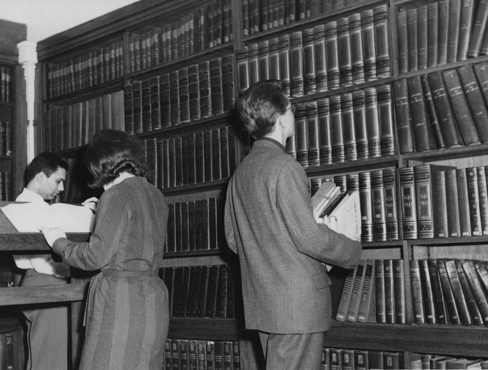 A man wearing a suit carrying a stack of books looks at a large bookshelf filled with encyclopedias. Behind him a woman is standing reading a book and another man browses the shelf in the distance.