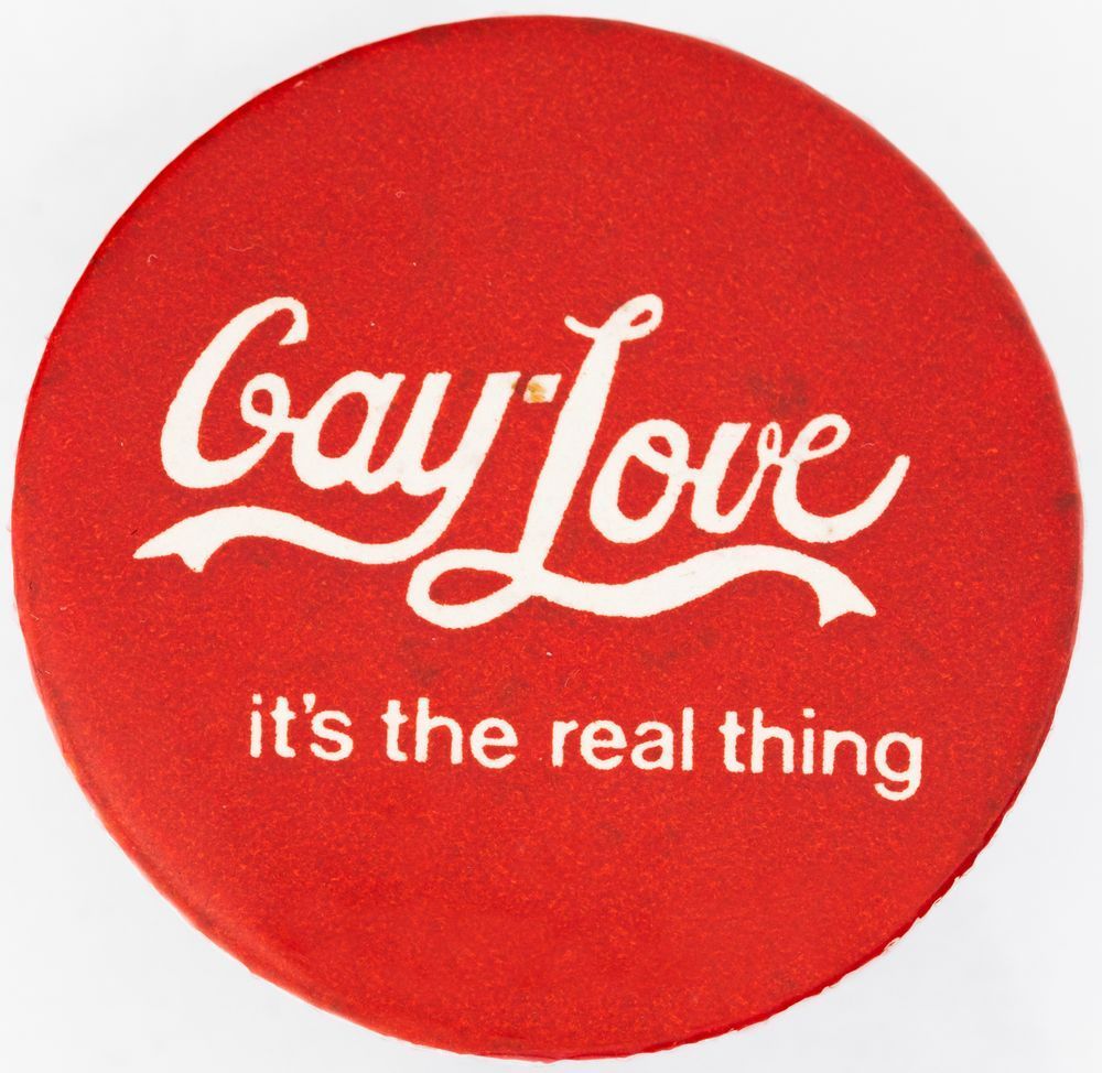 Red badge pin with white text resembling a Coca-Cola logo reading “Gay-Love it’s the real thing”
