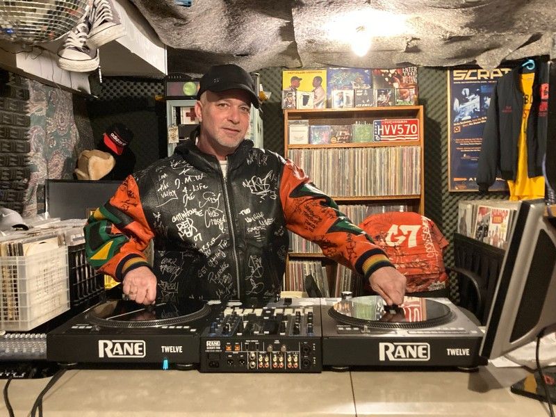 Scott Downer wears a leather jacket and stands in front of two turntables.