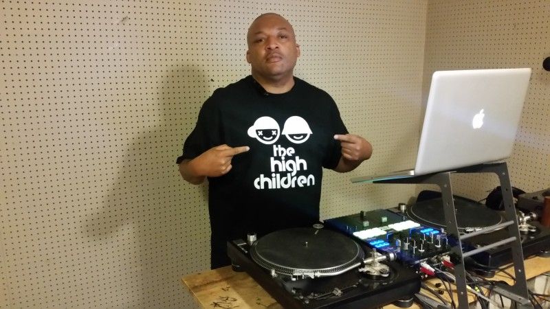 Will Higgs wears a black T-shirt of The High Children and stands in front of a laptop and turntables.