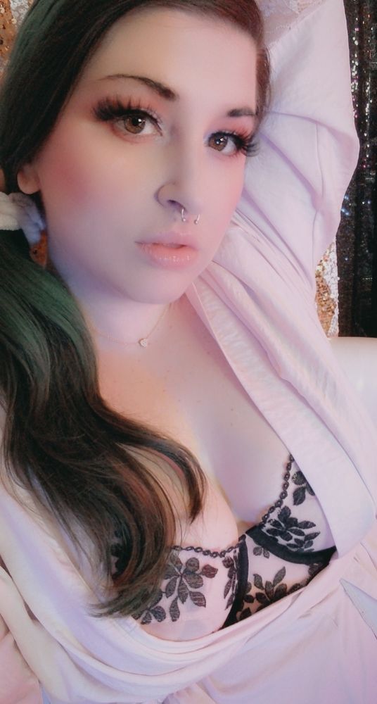 Findom Goddess Joules reclines gracefully, her confident gaze framed by dramatic lashes and soft, rosy makeup. She wears a delicate black floral lace bra under a flowing pale pink robe, complemented by a dainty heart-shaped necklace. The background features shimmering golden zig-zag patterns, enhancing the serene yet alluring atmosphere.