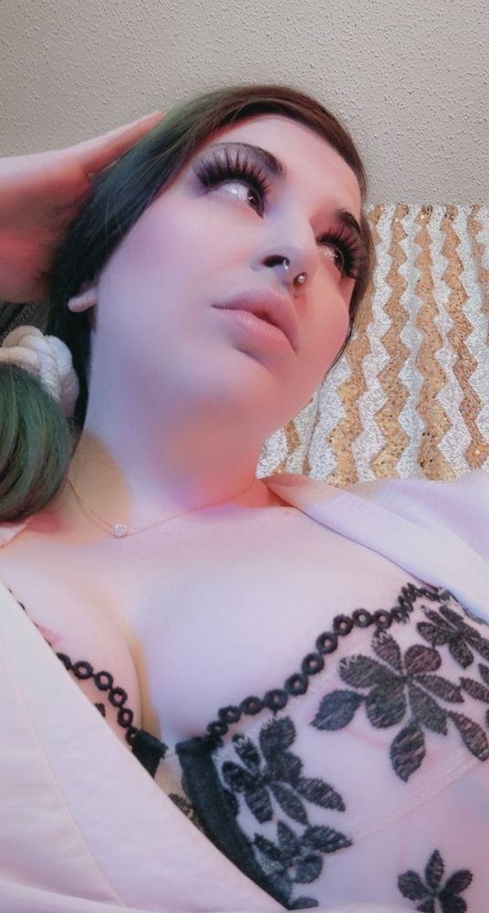 Hypnodomme Goddess Joules strikes a captivating pose, her head tilted upward to highlight her flawless features, accentuated by voluminous lashes and soft, natural makeup. She wears a sheer black floral lace bra beneath a pale pink robe, with a delicate necklace adding elegance. The golden zig-zag patterned backdrop adds a subtle touch of glamour. The camera is angled from below to remind you of your place.