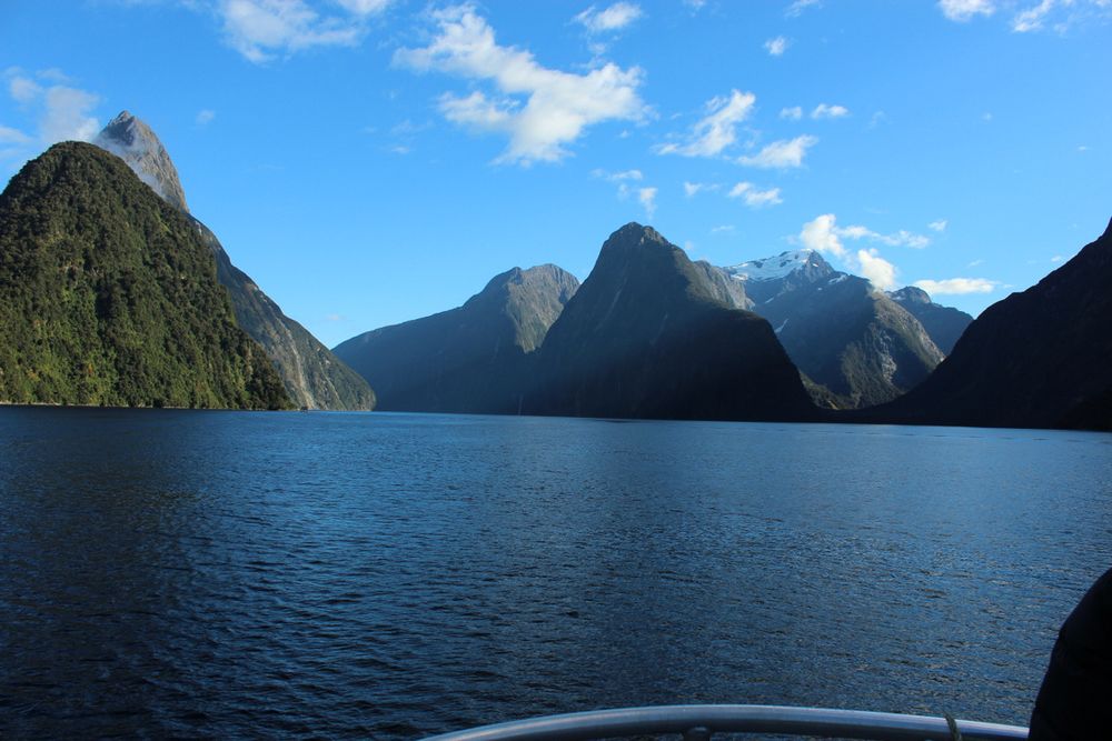 Milford Sound, NZ. View from a boat with blue water in front, mountainous fjords in front with snow caps on one, and blue skies with wispy white clouds.