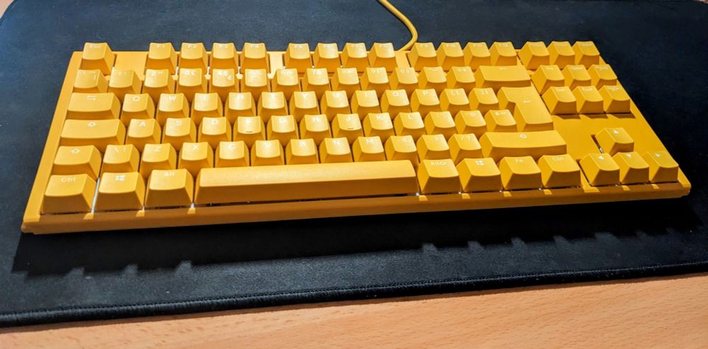 A yellow keyboard sitting on a black desk mat. It is a 75% mechanical keyboard with no numpad. The keys have white alphanumeric characters.