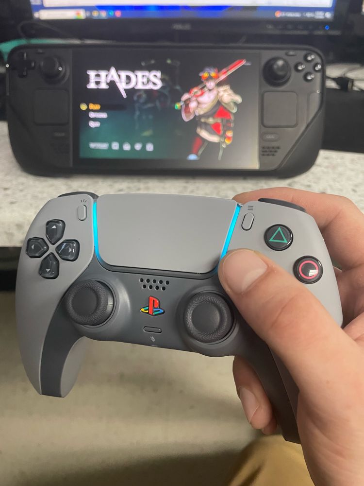 A hand holds a PS5 DualSense (30th Anniversary colors) in front of a Steam Deck playing Hades.