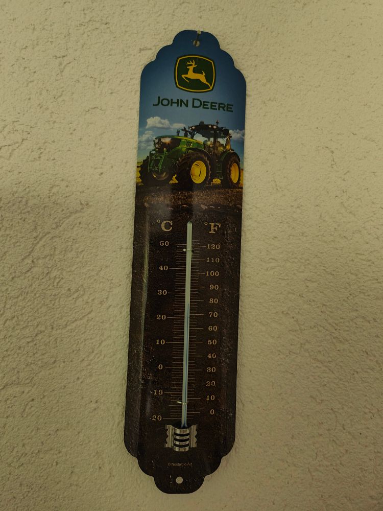 Analoges Thermometer vor der Rauputzwand. Zeigt 28 Grad Celsius.Hat aber auch eine 
Skala in Fahrenheit. Ausserdem ist eine Traktoren Marke auf dem Teil 
