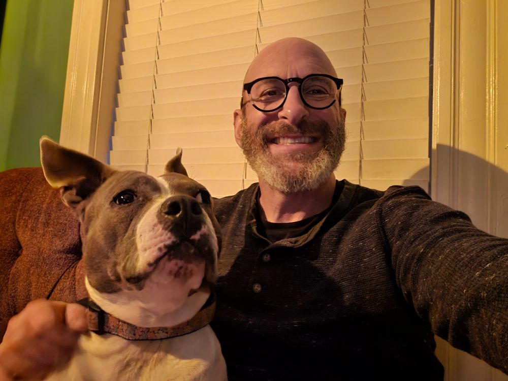 A blue and white American Bully sits next to a smiling man weating round black glasses