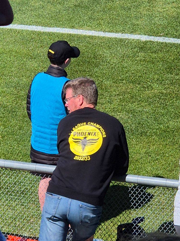 A man is wearing a shirt with a Wellington Phoenix logo and the words 'A-League Champions 2032/33'