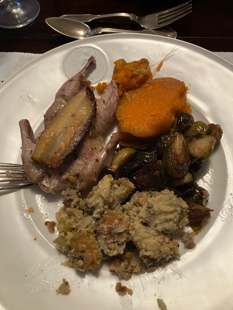 A china plate containing candied sweet potato, roasted Brussels sprouts, dressing, and a quail. 