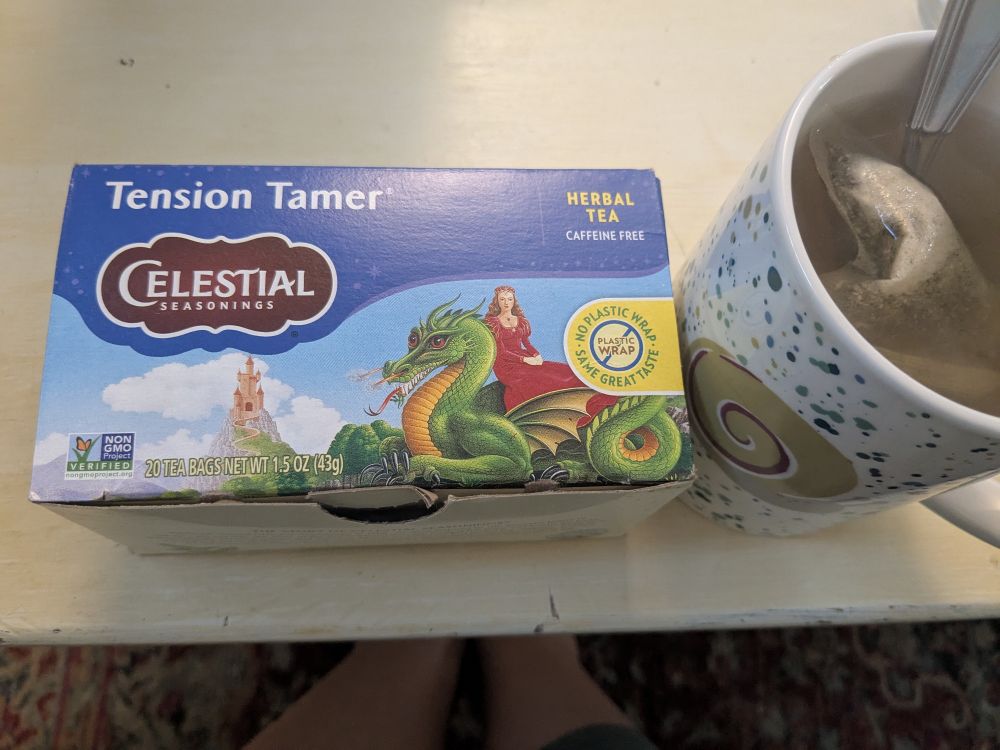 A box of celestial seasonings tension tamer tea, with an illustration of a green dragon and a woman wearing a red dress sitting on them. There is a castle in the background. There's a mug with a snail on it full of tea beside the box