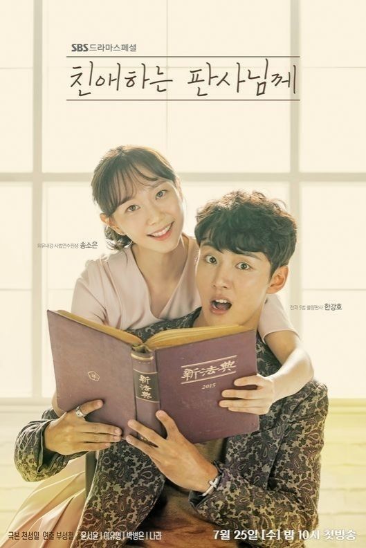 Um homem está sentado com um livro aberto nas mãos, por trás dele está um mulher sorrindo segurando uma das capas do livro.