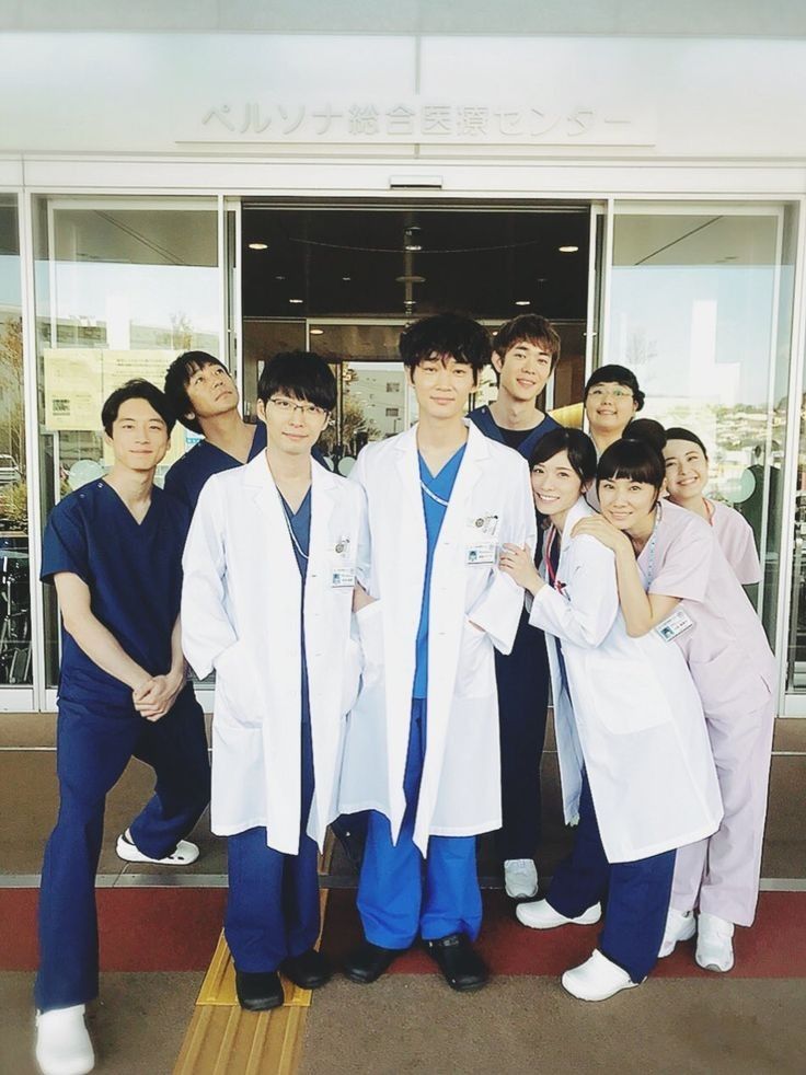 Os atores do drama Kounodori em uma foto juntos, há dois homens com jalecos, os médicos, também há  os médicos residentes de azul, e do lado deles pessoas da equipe de enfermagem, no total há 9 pessoas na foto. 