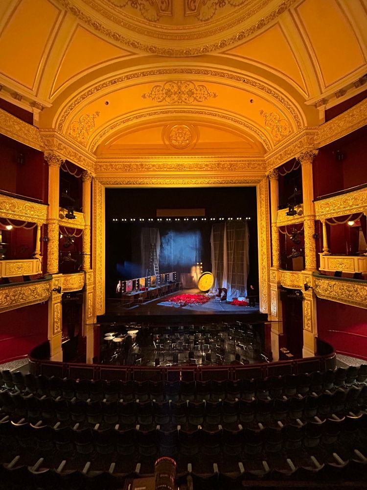 The interior of the Theatre Royal Glasgow, lit in gold.
On the stage is a back-stage set with a pile of deep red roses, old dressing table mirrors, tall masking flats covered in a voile, and a large golden oversized clock. 