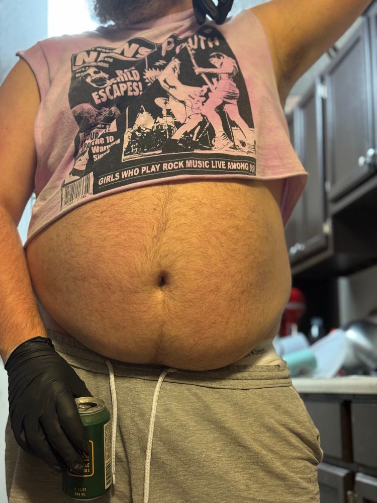 A guy standing wearing a crop top, tiny shorts, black gloves, and holding a bear while showing off his belly