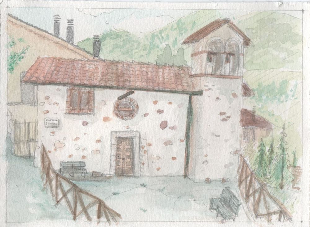 A white church with bricks and stone of different colours and an orange roof. The building is surrounded by fences made with branches. There are other buildings around and hills in the background.