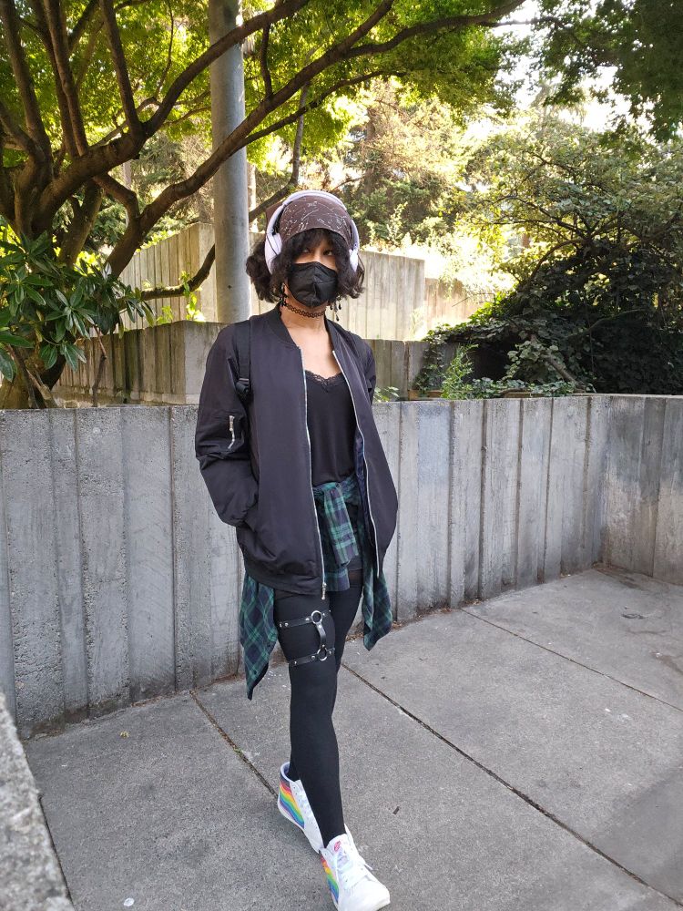 A pic of me looking to the side in a park wearing a black bandana, lavender headphones, black gothic cross earrings, black mask and choker, black bomber jacket and black cami top. I have a green flannel tied around my waist and black tights with a dual connection leg harness. On my feet, I have white with rainbow accents hi-top converse!