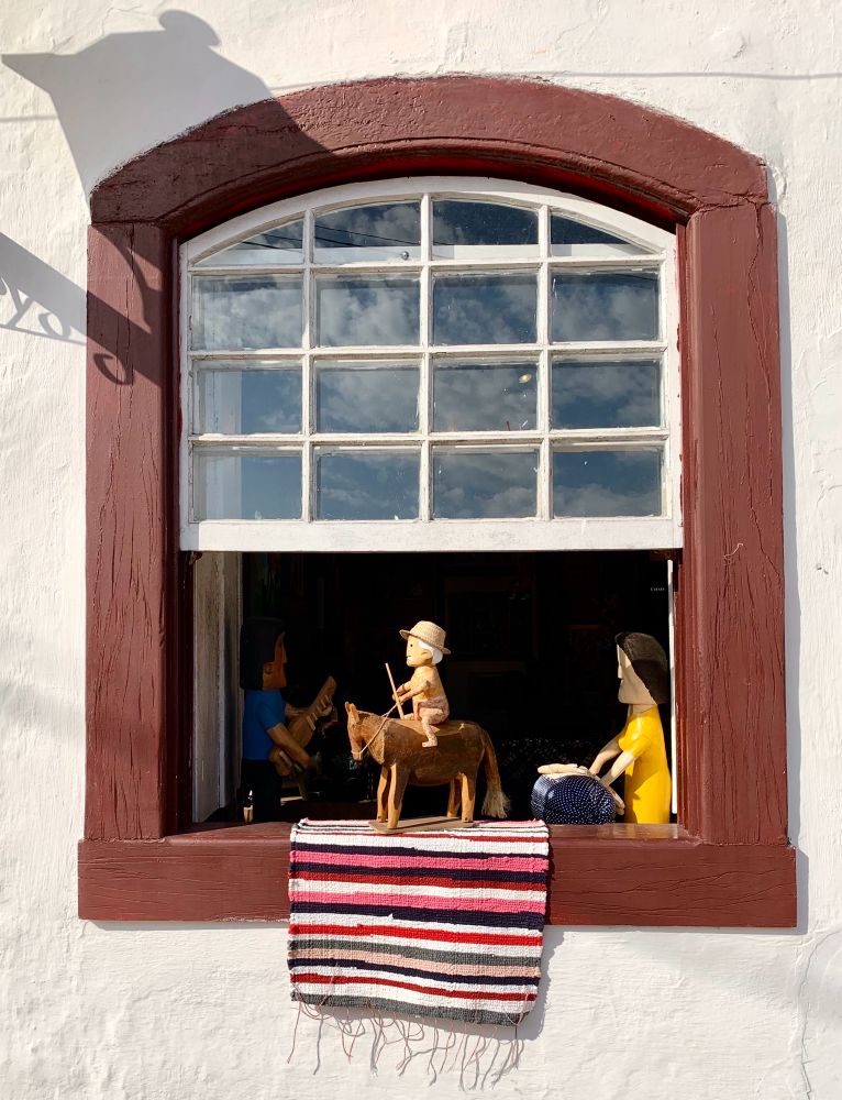 Window framed with brown wood arch in white stucco building. Wooden toys are placed on or near the windowsill. One is a man on a brown horse placed on a small brightly colored striped rug. Upper half of window shows the raised window with 12 rectangular windows framed in white. 