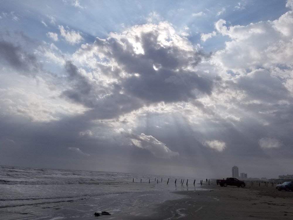 A picture of the beach in Galveston, Texas. A large fluffy cloud is blocking the sun causing fingers of light and shadow to reach down from the sky.