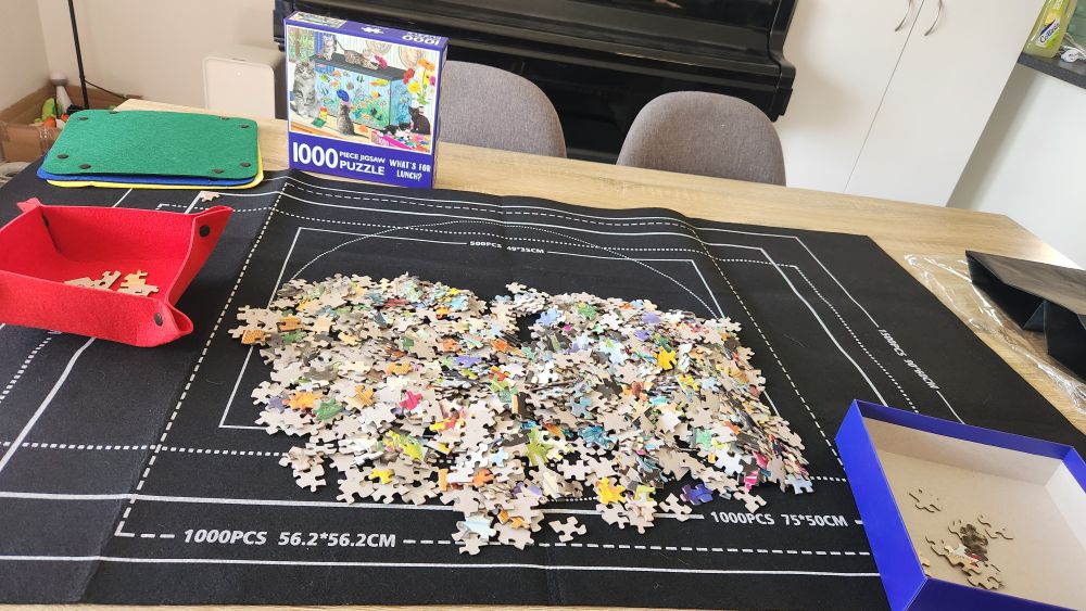 A pile of jigsaw puzzle pieces plopped in the middle of a jigsaw mat, with some sorting trays to the sides 