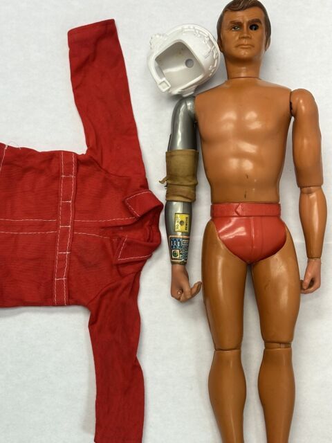 photo of the "Six Million Dollar Man" doll with his tiny red onesie to one side, his robotic arm and extremely tight red belted undies on an otherwise naked, one-eyed figure, and an astronaut helmet to one side.