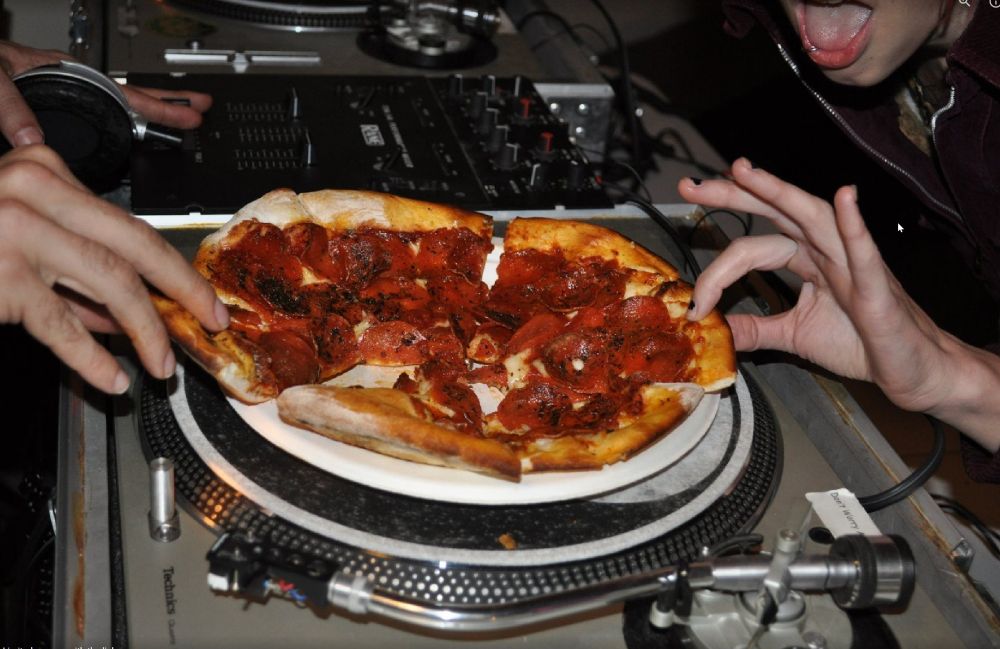 close up of pizza on a turntable. I am reaching to take a slice and so is a person on the right, her hand visible and part of her face with open mouth