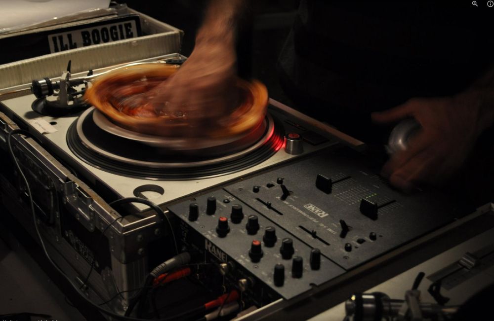 close up of turntables and mixer, with pizza on a plate on one turntable. I am DJing the pizza on my right, headphone in my left hand and fingers over the mixer. The pizza is spinning so fast it is blurred in the photo