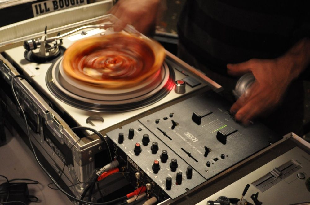 close up of turntables and mixer, with pizza on a plate on one turntable. I am DJing the pizza on my right, headphone in my left hand and fingers over the mixer. The pizza and my hand are both blurred from fast motion 