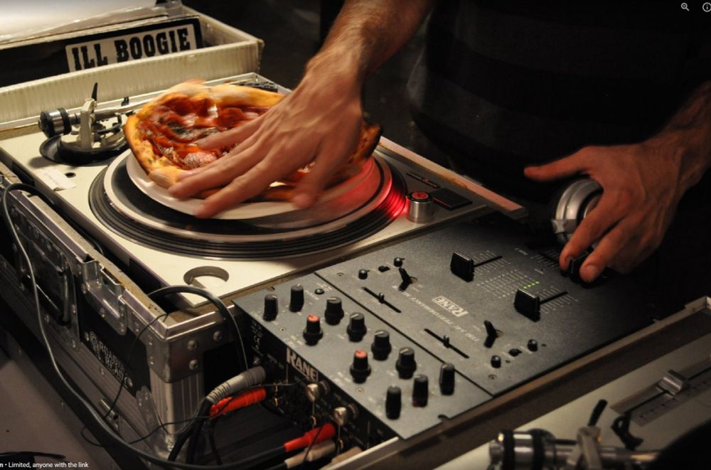 close up of turntables and mixer, with pizza on a plate on one turntable. I am DJing the pizza on my right, headphone in my left hand and fingers over the mixer
