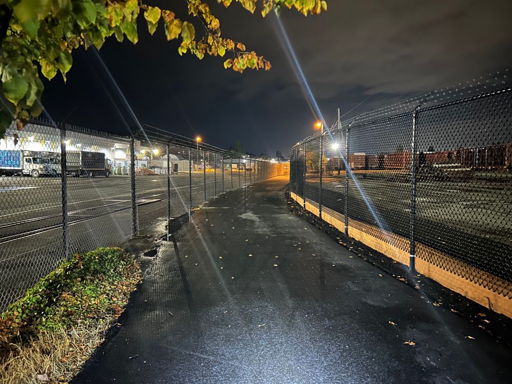 A newly paved trail at night that is flat and spacious. There are fences in both sides of the trail as this runs between some industrial buildings and a rail yard. 