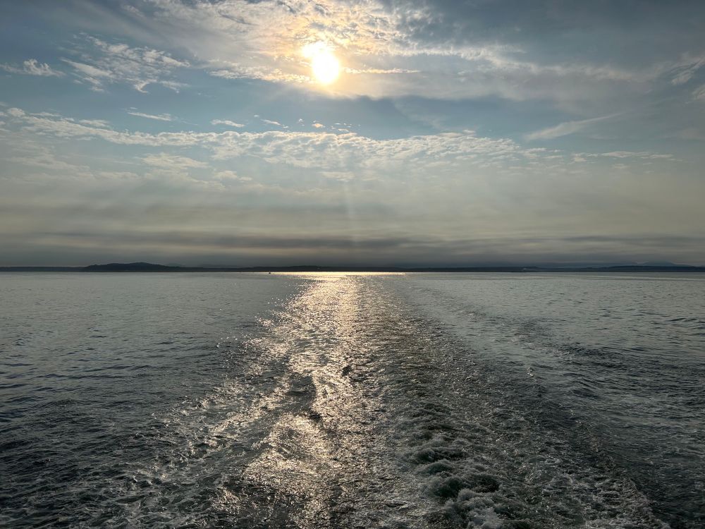 A picture of some waves in the ocean with the sun in the distance. It’s not quite sunset, but the sun is approaching the horizon and starting to turn the yellow orange color of sunsets. There’s a smattering of clouds in the sky, with the sun slightly behind a thinner cloud. 
