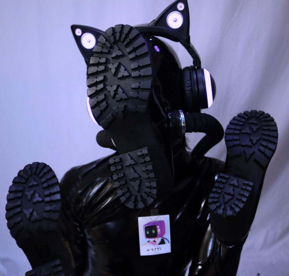 Boot drone, covered in shiny black latex and wearing boots over its hands, and face, and wearing cat ear heaphones. The drone has a polaroid of its previous identity as a tv-headed maid taped to its chest, with it's drone ID written on it. The boot drone is waving at you with both boots :3