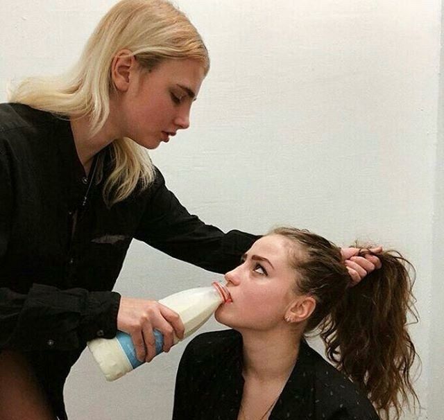 A blond woman holds a bottle of milk in one hand and the ponytail of a brunette woman in the other. The blond woman is encouraging the brunette to drink the milk as the brunette looks up towards the blonde woman. 