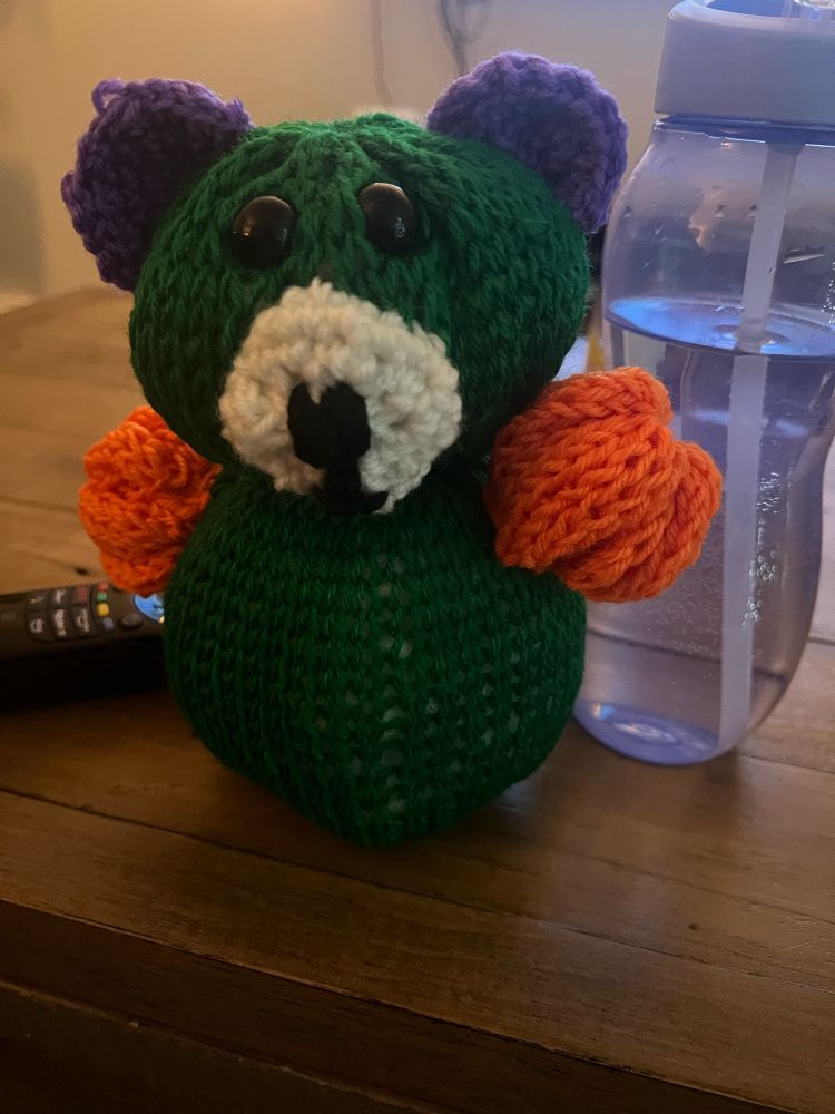 A knitted teddy bear featuring a green body, white muzzle, purple ears, orange arms but no legs yet. 