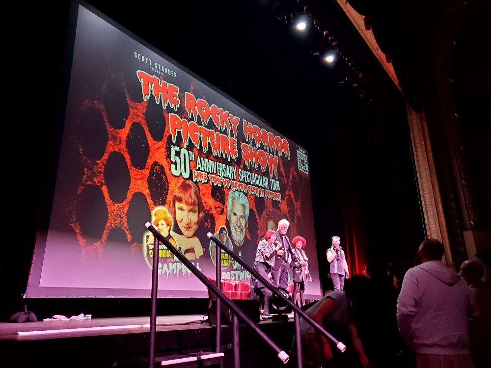 On stage are Nell Campbell (Columbia), Barry Bostwick (Brad), and Patricia Quinn (Magenta), along with an event emcee. The screen behind them reads "The Rocky Horror Picture Show 50th Anniversary Spectacular Tour."