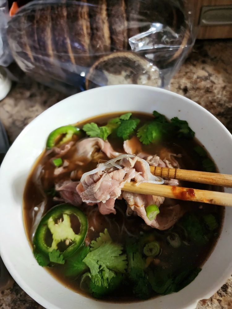 A bowl of pho bo, a Vietnamese noodle soup with rice noodles, thinly sliced steak, and a rich beef broth, garnished with scallion, cilantro, and jalapeño. A pair of chopsticks lifts a bite of noodles and steak toward the viewer.