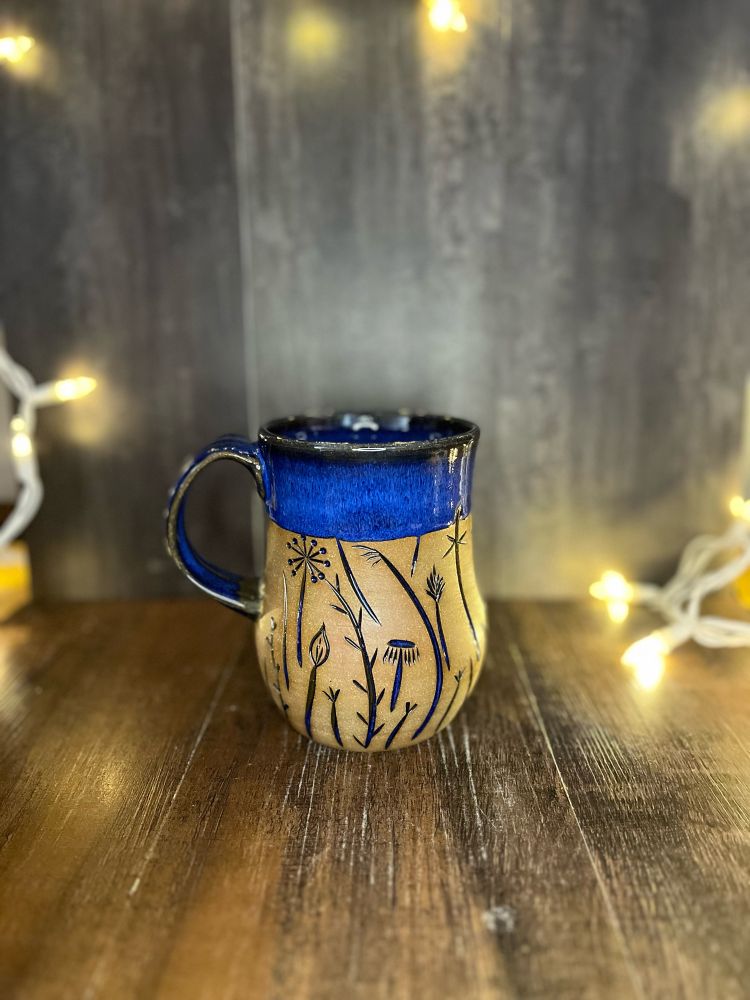 A carved mug with a cobalt blue glaze and carving that look like a summer field of plants and insects. There are white twinkle lights blurred in the background, giving it a magical look. 