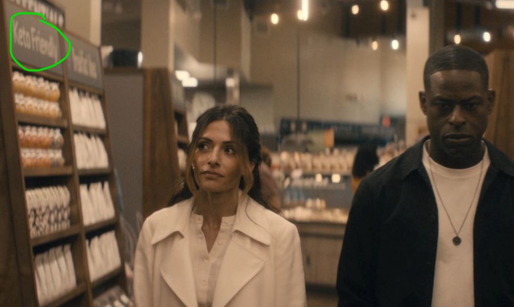 Screenshot from Paradise. Sarah Shahi and Sterling K. Brown are seen from the front walking down a grocery store aisle. In the upper left corner is a shelf labeled “Keto Friendly”